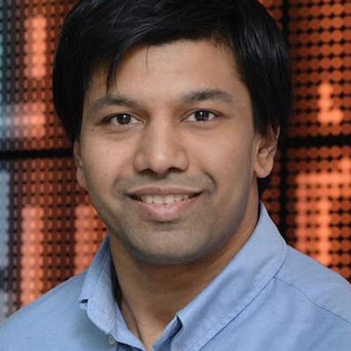  A headshot shows a smiling man with black hair wearing a light blue button-down shirt, with a blurred, orange and brown patterned background.